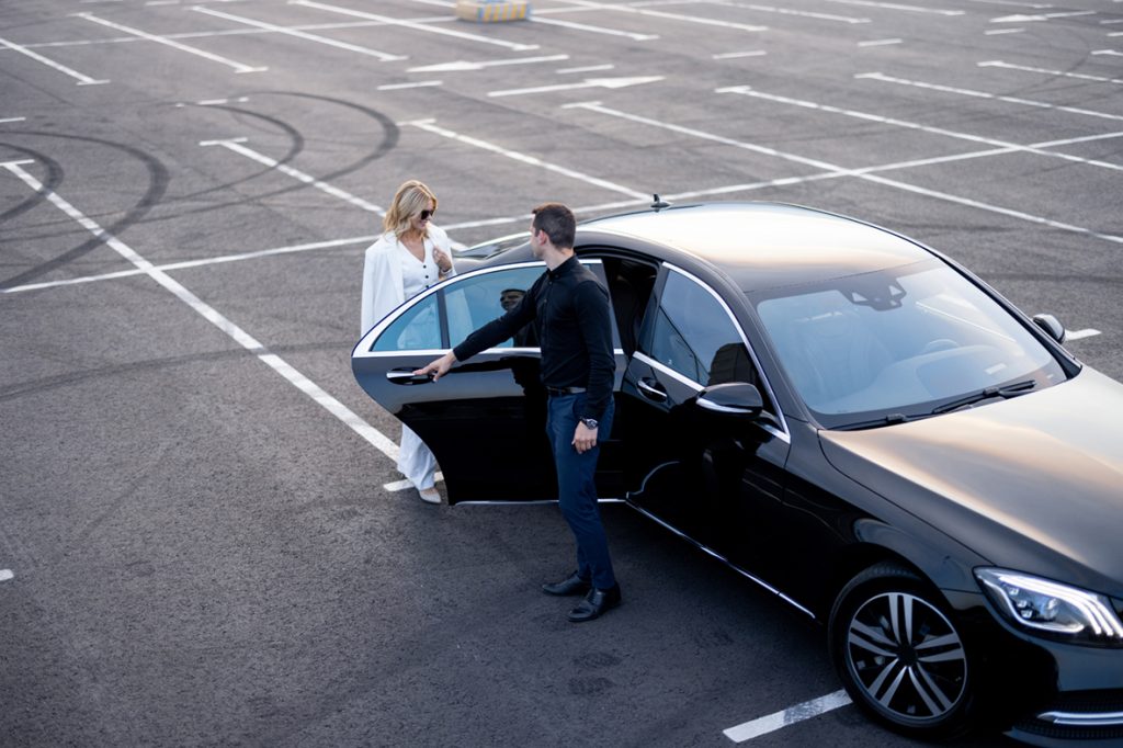 Wir sehen eine schwarze Limousine, eine Frau mit weißer Kleidung beim Einstieg in das Fahrzeug und einen Fahrer, der die Tür zum Einstieg aufhält.